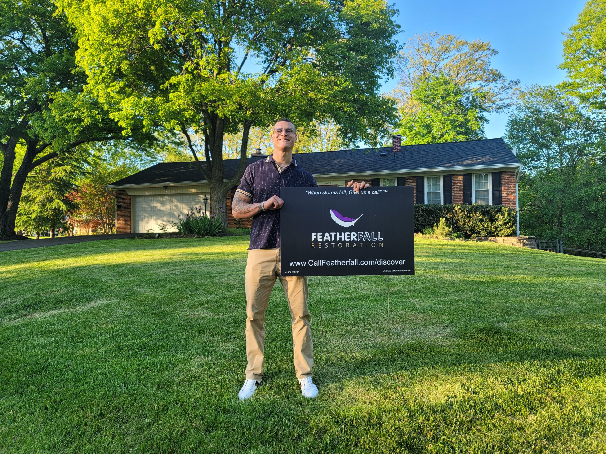 Smiling man standing on a lawn holding a Featherfall Restoration yard sign in front of a brick house with a recent roofing and siding replacement
