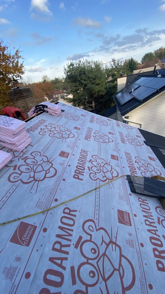 Roof covered with Owens Corning ProArmor underlayment, stacks of roofing materials, and solar panels visible on a neighboring house during roof replacement