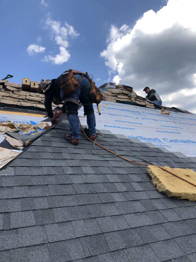 Two workers installing asphalt shingles on a roof for a roof replacement, surrounded by roofing materials and underlayment, beneath a partly cloudy sky