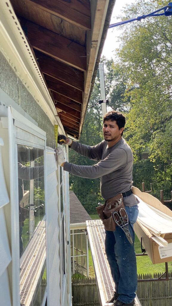 Construction worker standing on scaffolding, measuring siding near a window during siding replacement beneath exposed roof eaves