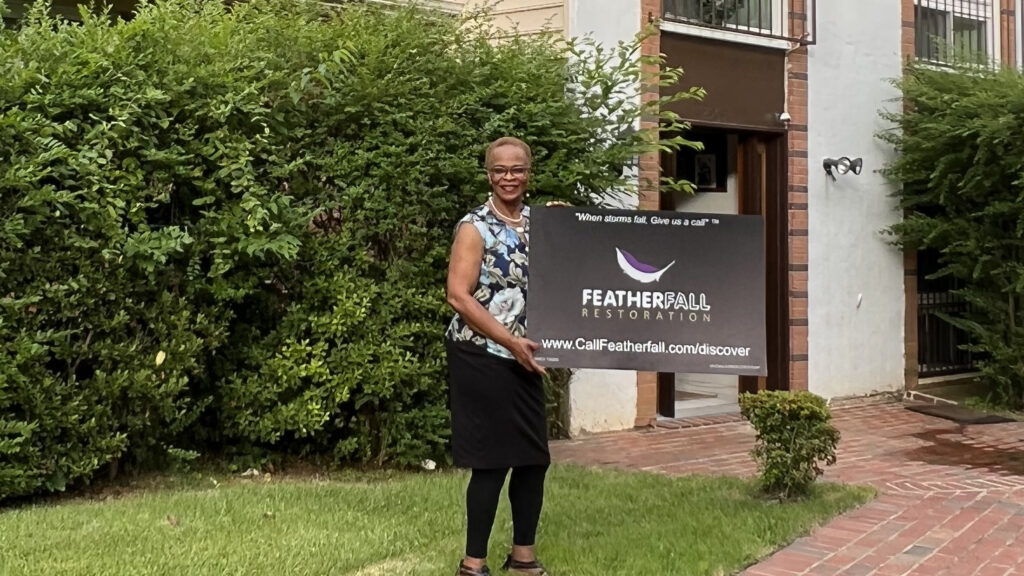 Smiling homeowner holding a Featherfall Restoration sign in front of a brick walkway and entryway surrounded by greenery after her siding replacement