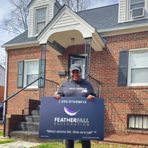 Smiling homeowner holding a Featherfall Restoration yard sign in front of a brick house with dormer windows and black shutters after a roof replacement