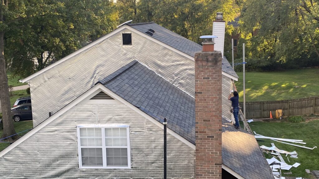 Rear view of a house during siding replacement with exposed insulation wrap, scaffolding, and a worker installing siding near the chimney